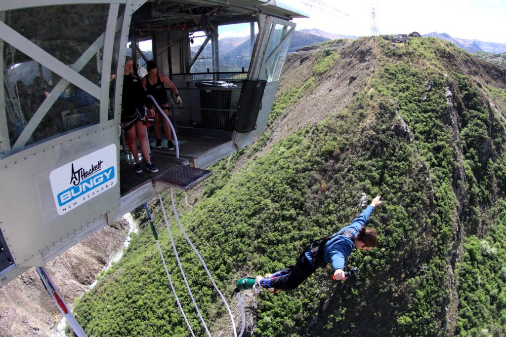 Person leaping from the AJ Hackett Bungy platform high above a canyon in Queenstown, New Zealand, with mountain scenery behind