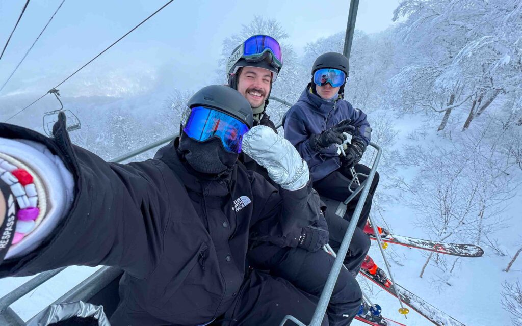 Three friends taking a selfie on a ski lift at Nozawa Onsen resort in Japan, surrounded by snow-covered trees