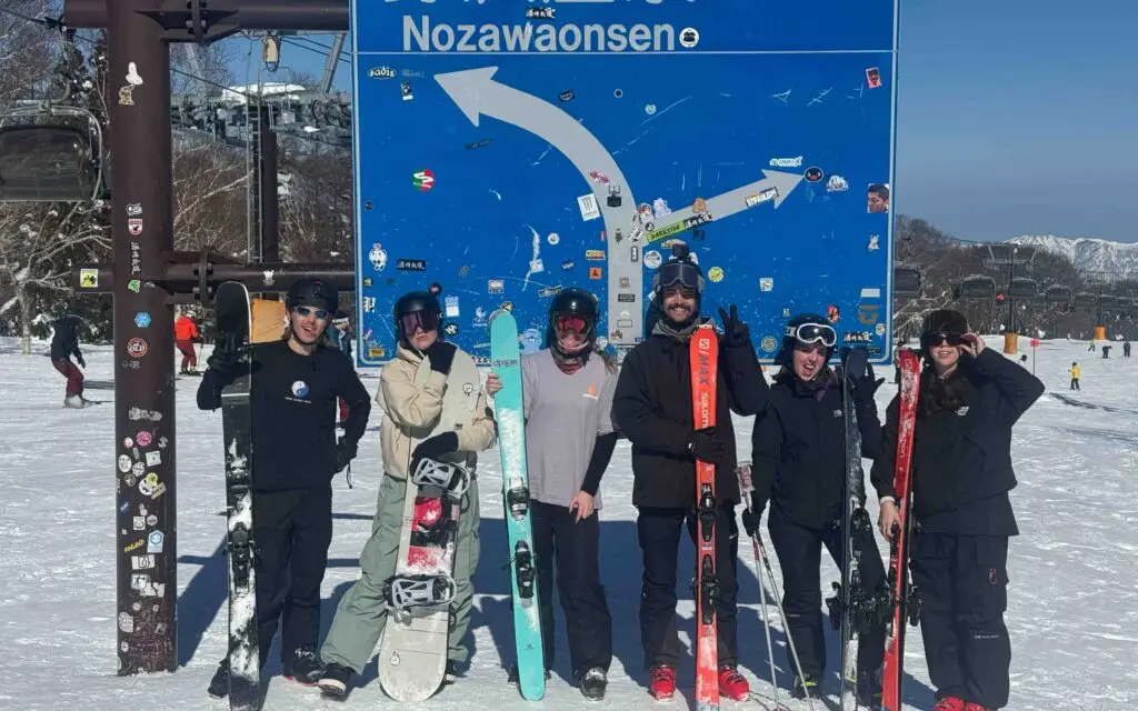 Group of skiers and snowboarders posing in front of the iconic blue Nozawa Onsen resort sign on a sunny day in Japan