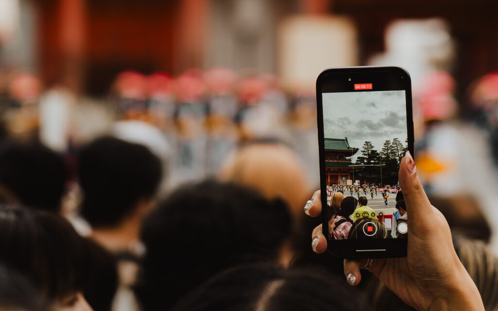 Hand holding an iPhone filming the Jidai Matsuri festival procession at Heian Shrine in Kyoto, Japan, through a crowd of spectators