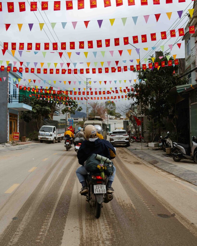 Two people riding a motorbike through a Vietnamese town street decorated with colourful bunting and red flags on the Ha Giang Loop, Vietnam