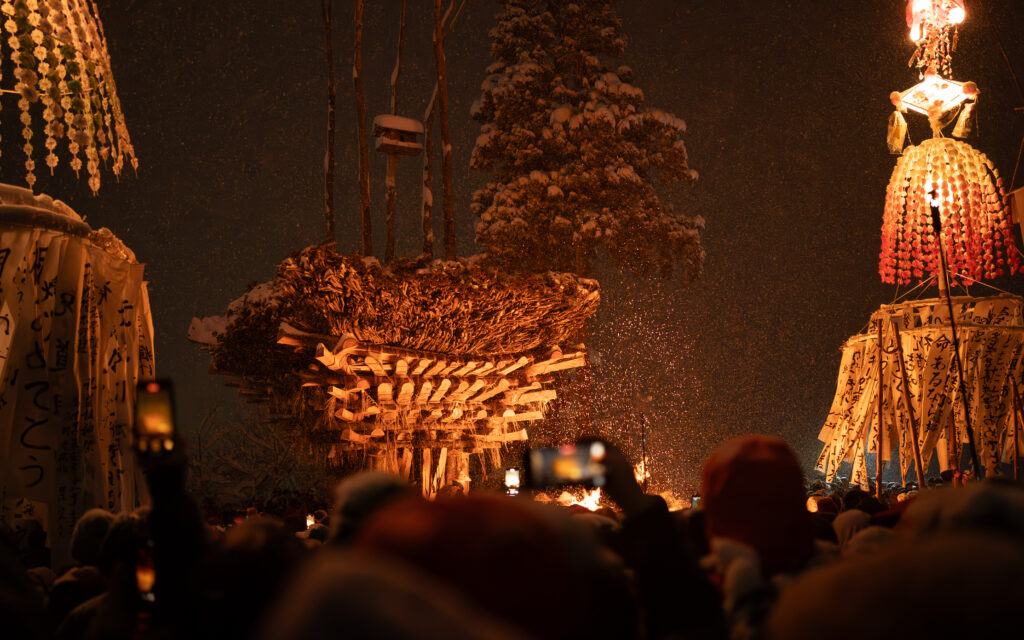 Large wooden structure glowing with fire at the Nozawa Onsen Dosojin Fire Festival, surrounded by crowds and traditional Japanese paper banners