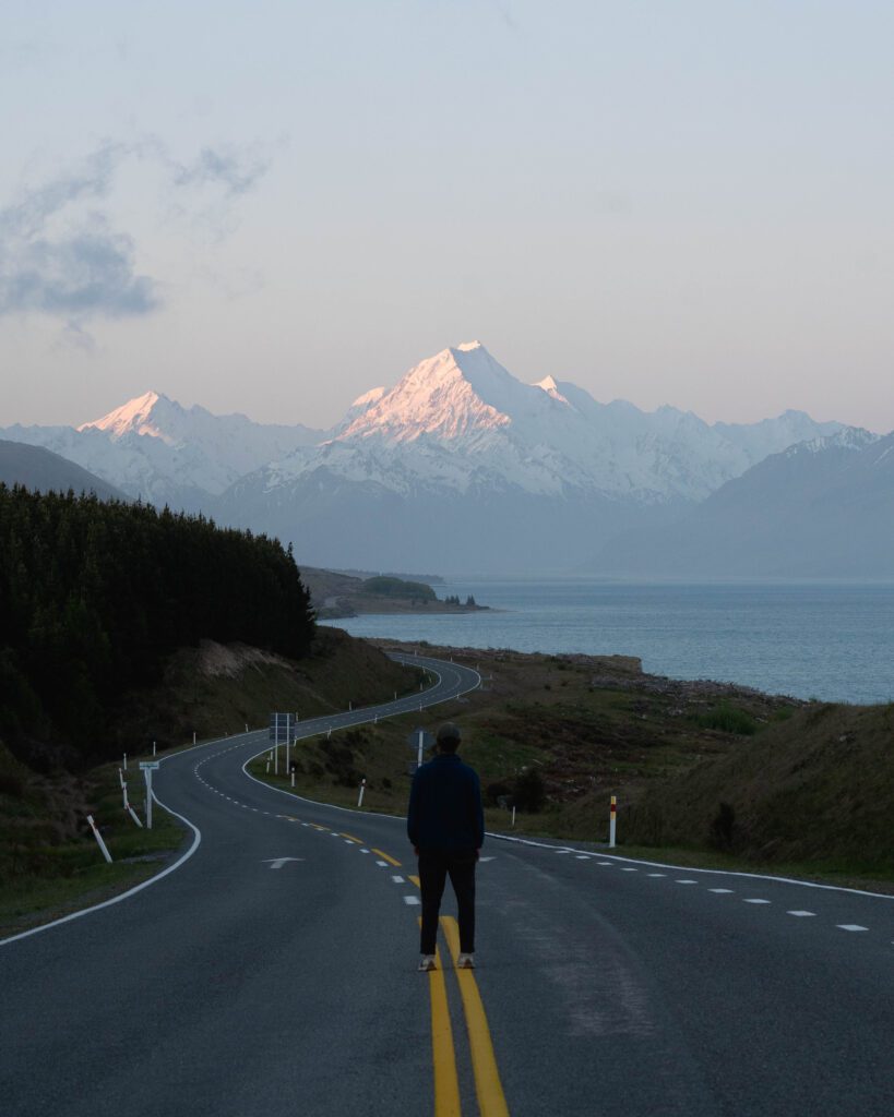 Person standing on a winding road beside Lake Pukaki at dusk, facing snow-capped Aoraki Mount Cook in New Zealan