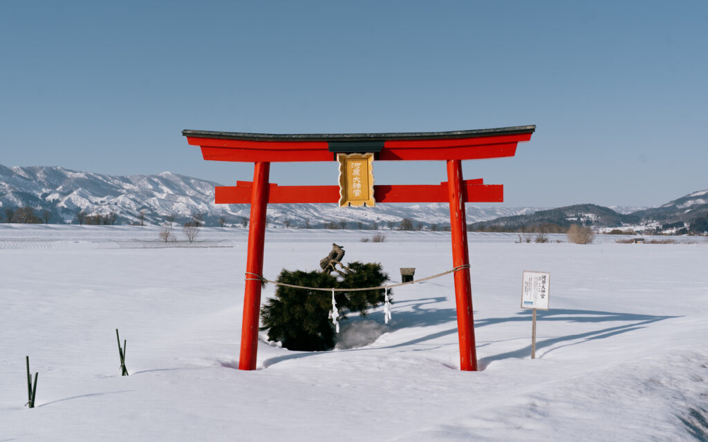 Bright red torii gate standing alone in a vast snowy field near Nozawa Onsen, with snow-capped mountains behind under a clear blue sky