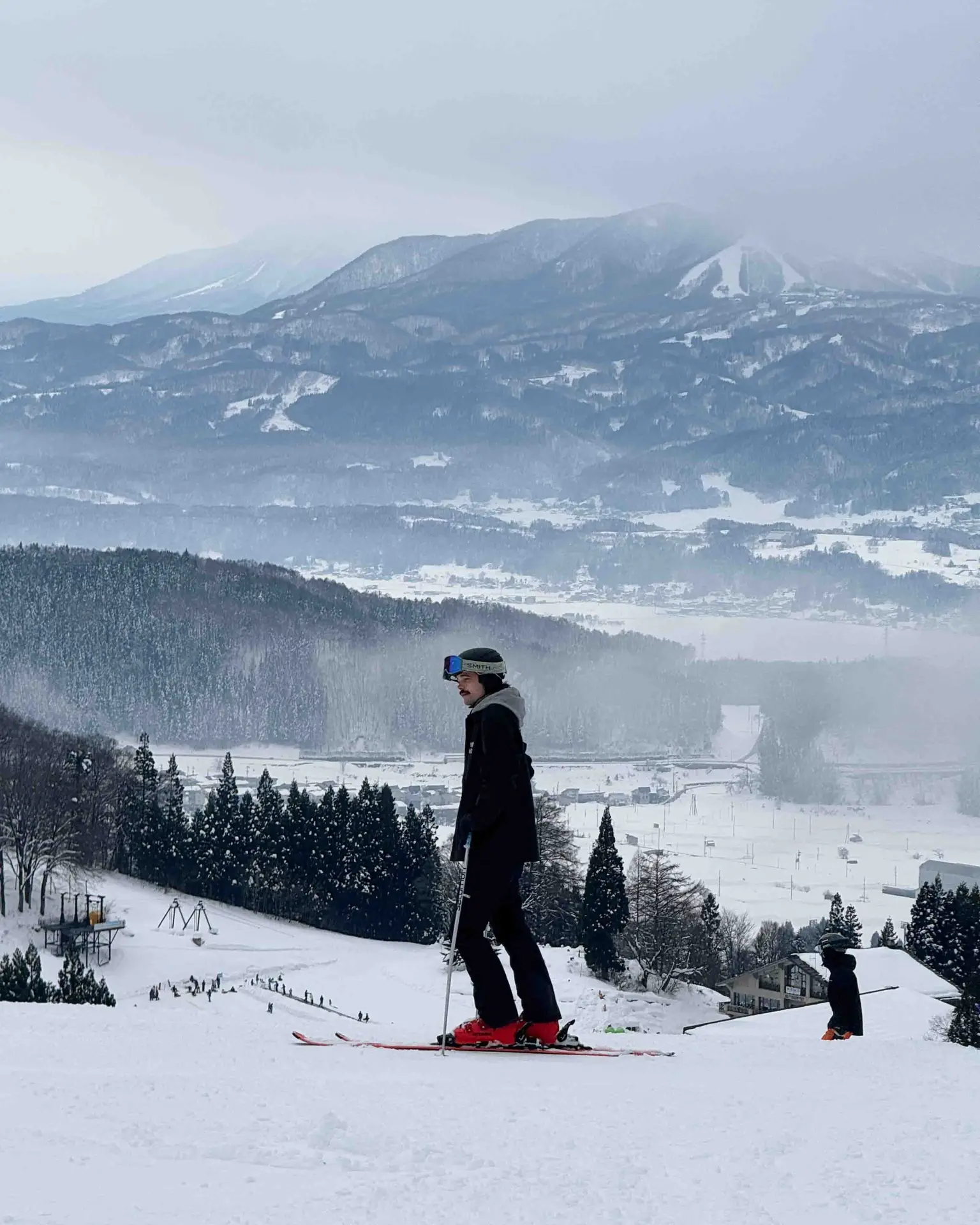 Skier pausing on the slopes at Nozawa Onsen with the snow-covered village and misty mountains visible below, Japan