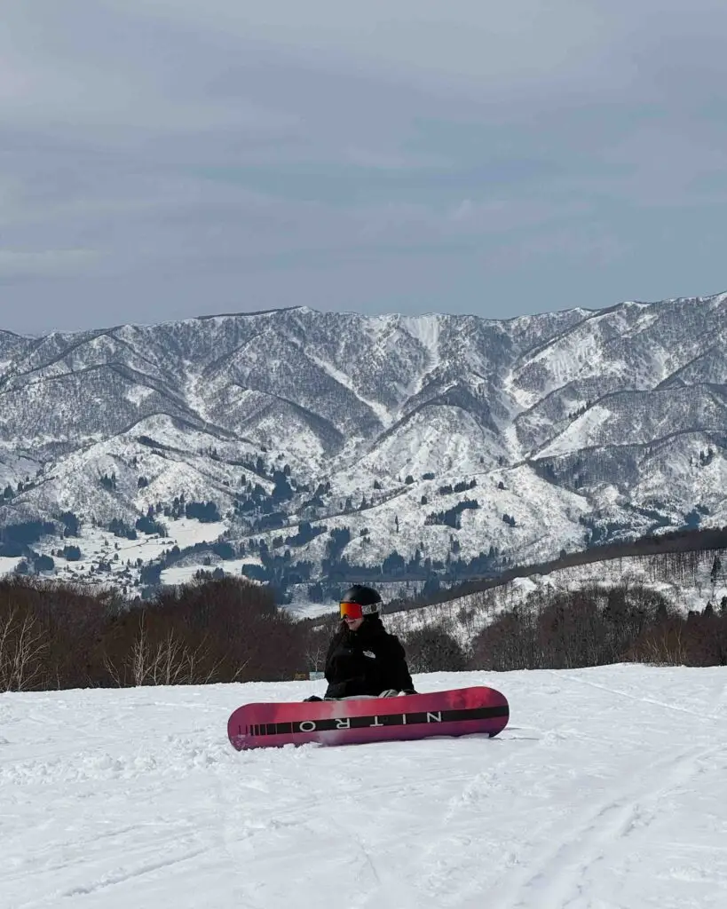 Snowboarder sitting on the slope at Nozawa Onsen with a Nitro board, with sweeping views of snow-covered Japanese mountains behind