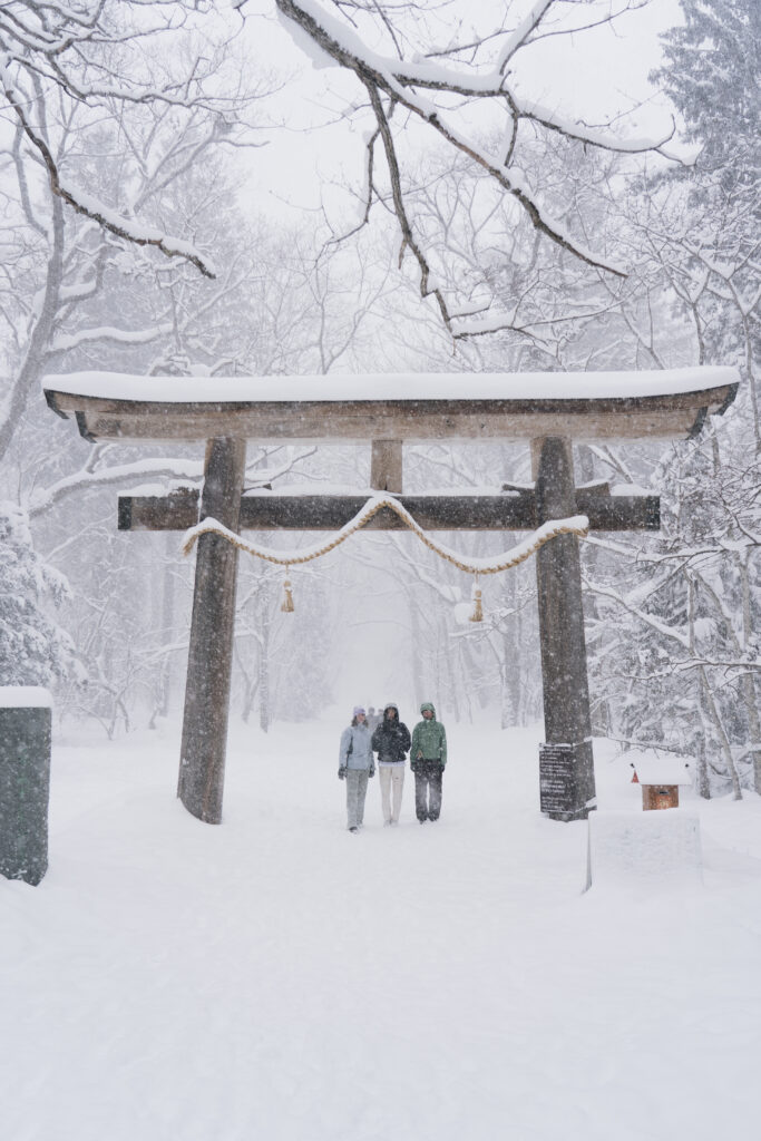 Three people standing beneath a stone torii gate covered in snow during a heavy snowfall near Nozawa Onsen, Japan
