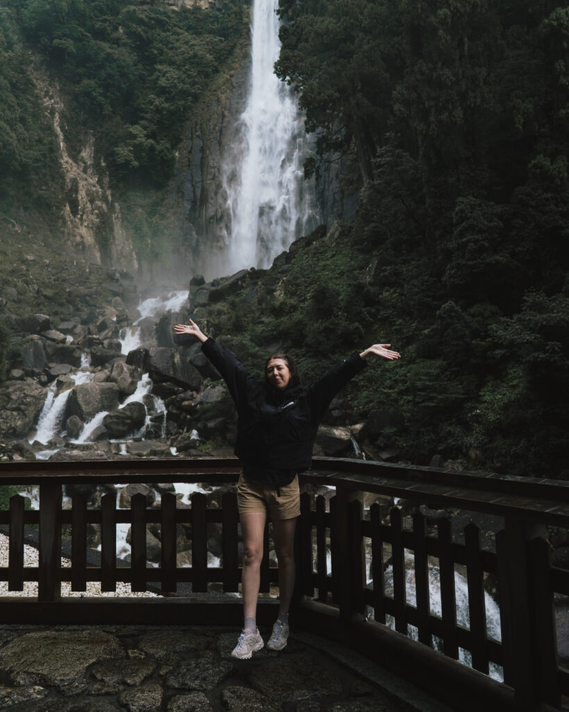 Woman with arms raised in front of Nachi Falls, Japan's tallest waterfall, in a remote forested location on the Kumano Kodo trail