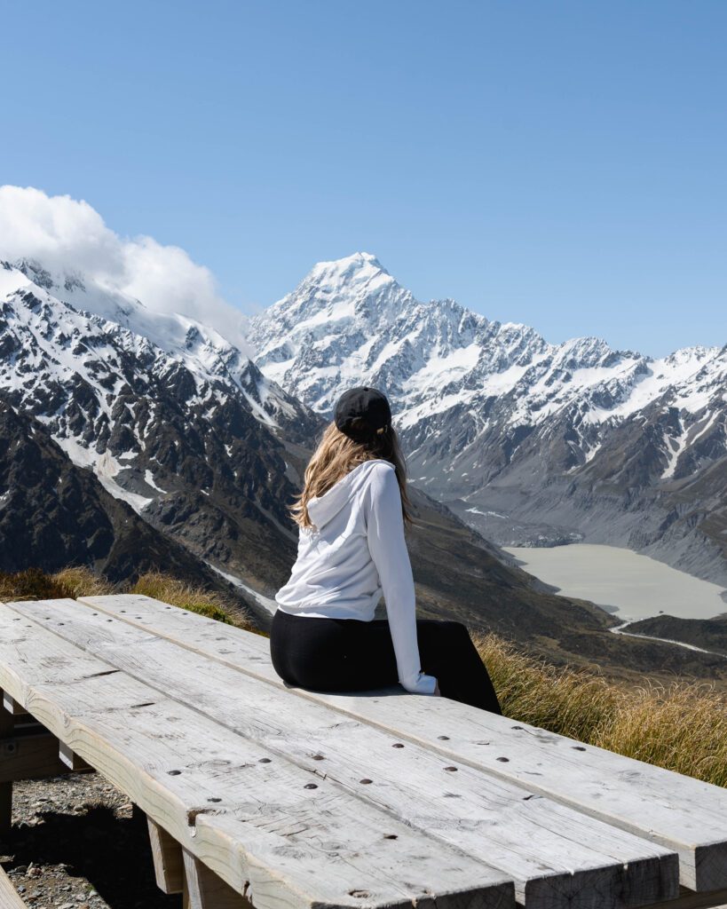 Woman sitting on a wooden viewpoint bench gazing at snow-capped Aoraki Mount Cook and Hooker Valley glacier, New Zealand