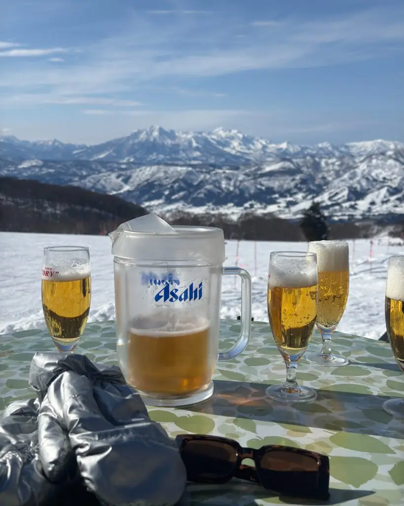 Asahi beer pitcher and glasses on an outdoor table at Nozawa Onsen ski resort with snow-covered mountains in the background, Japan