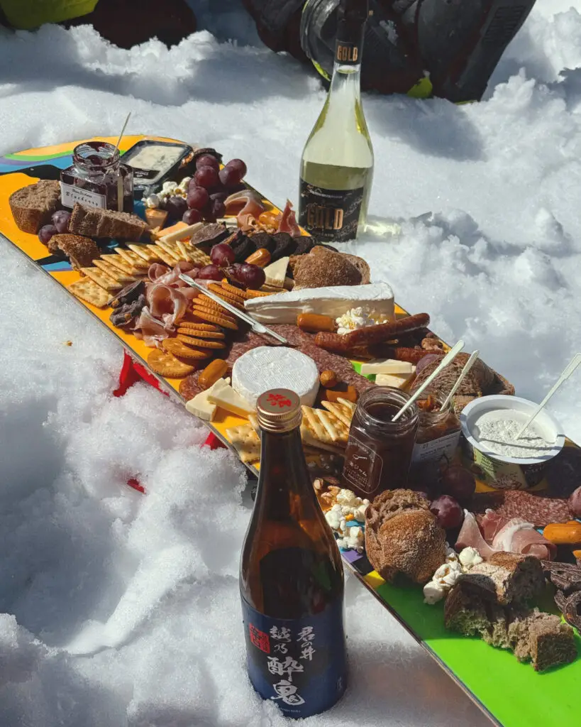 Charcuterie board loaded with cheese, crackers, meats and grapes laid out on a snowboard in the snow at Nozawa Onsen, with sake and wine bottles alongside