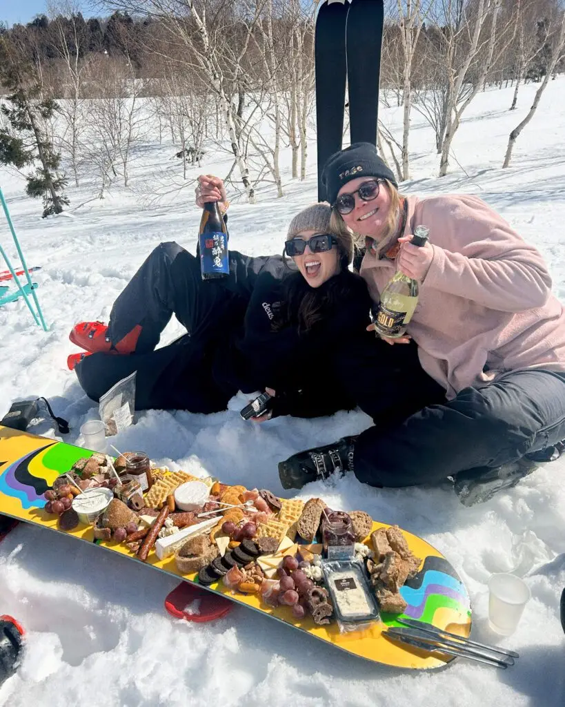 Two skiers laughing and holding sake and wine bottles beside a charcuterie board laid out on a snowboard in the snow at Nozawa Onsen, Japan