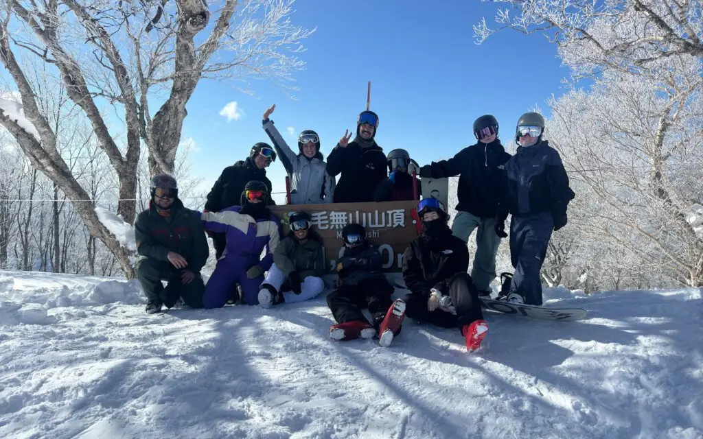 Large group of skiers and snowboarders posing at the summit sign of Kenashi mountain at Nozawa Onsen resort, Japan, on a clear blue sky day