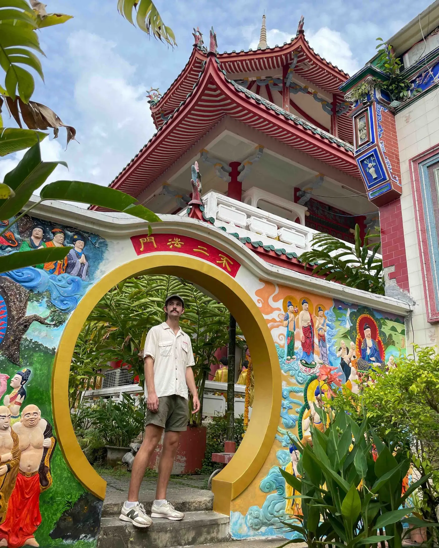 Man standing inside a circular golden gateway at a colourful Chinese Buddhist temple in Penang, Malaysia, surrounded by painted murals and traditional architecture. Best travel insurance for backpackers