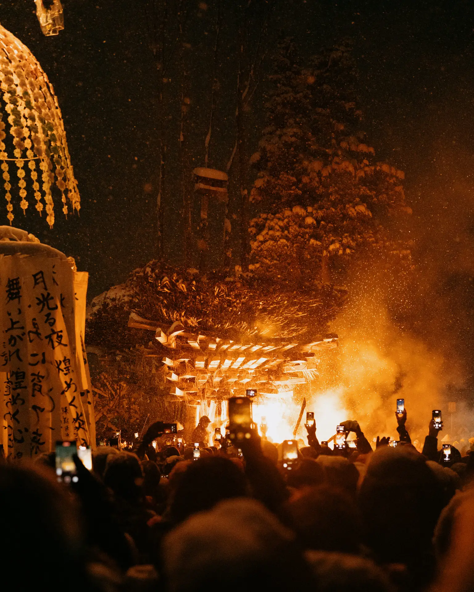 Crowd holding up phones to film the glowing shaden pyre at night during the Dosojin Fire Festival in Nozawa Onsen, with smoke rising and paper banners on the left