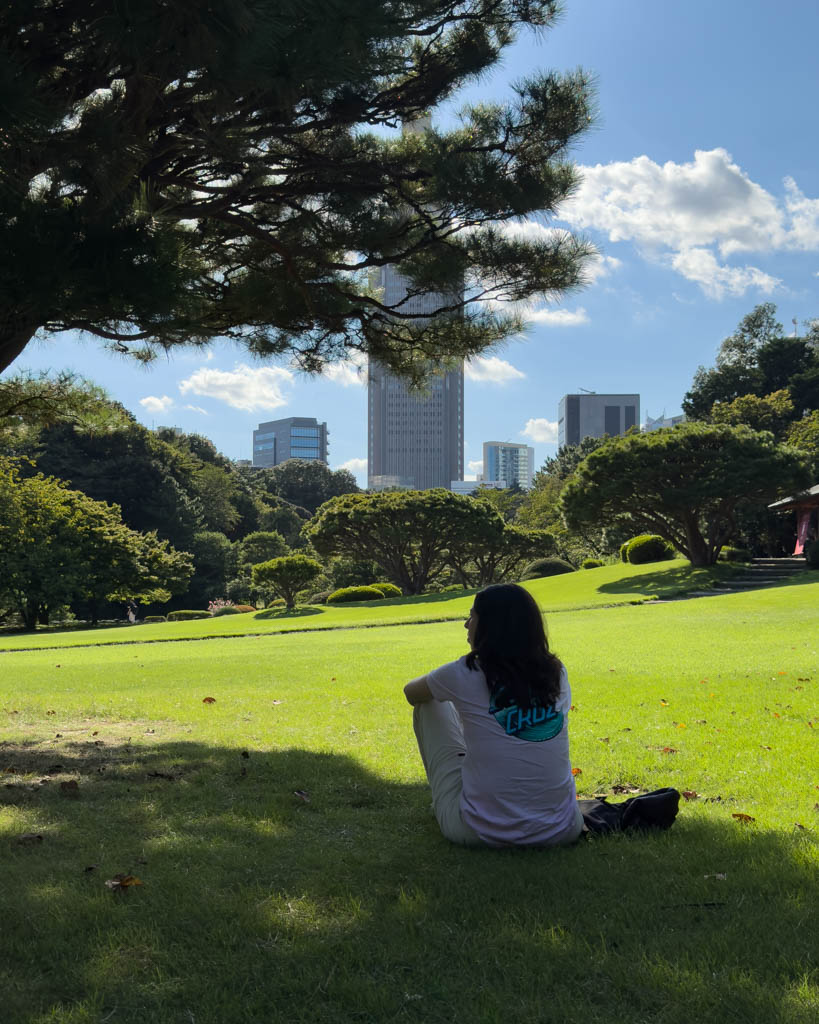 Visitor relaxing in the shade of a pine tree at Shinjuku Gyoen with a skyscraper backdrop in summer