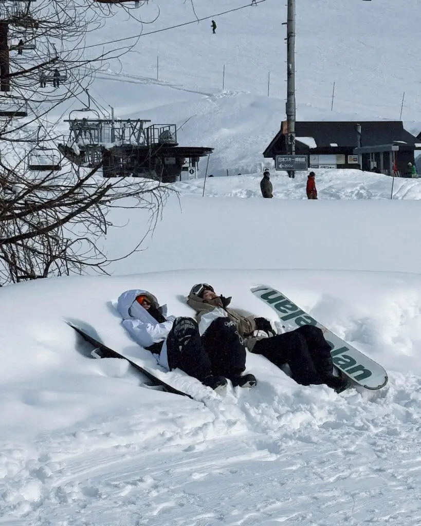 Two snowboarders sitting in deep snow on the slopes at Nozawa Onsen ski resort, Japan, with the chairlift and mountain buildings in the background