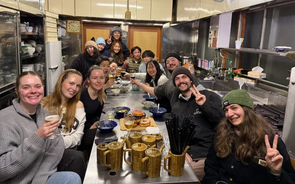Seasonal workers and Japanese staff sharing a meal together in the kitchen of Tatsumi restaurant in Nozawa Onsen, Japan, raising glasses and smiling