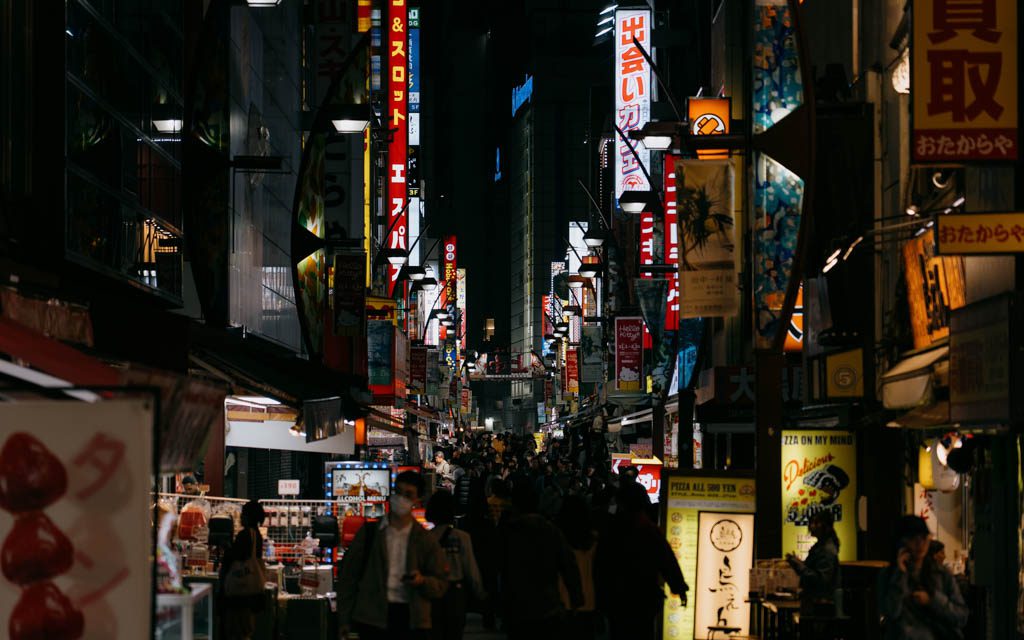 Crowds walking a narrow backstreet lined with illuminated signage near Ueno at night