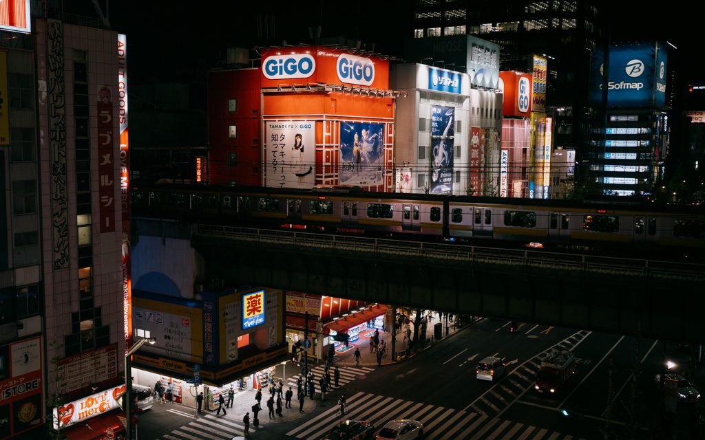 Neon-lit Akihabara street at night with a train passing overhead and GiGO arcade signage