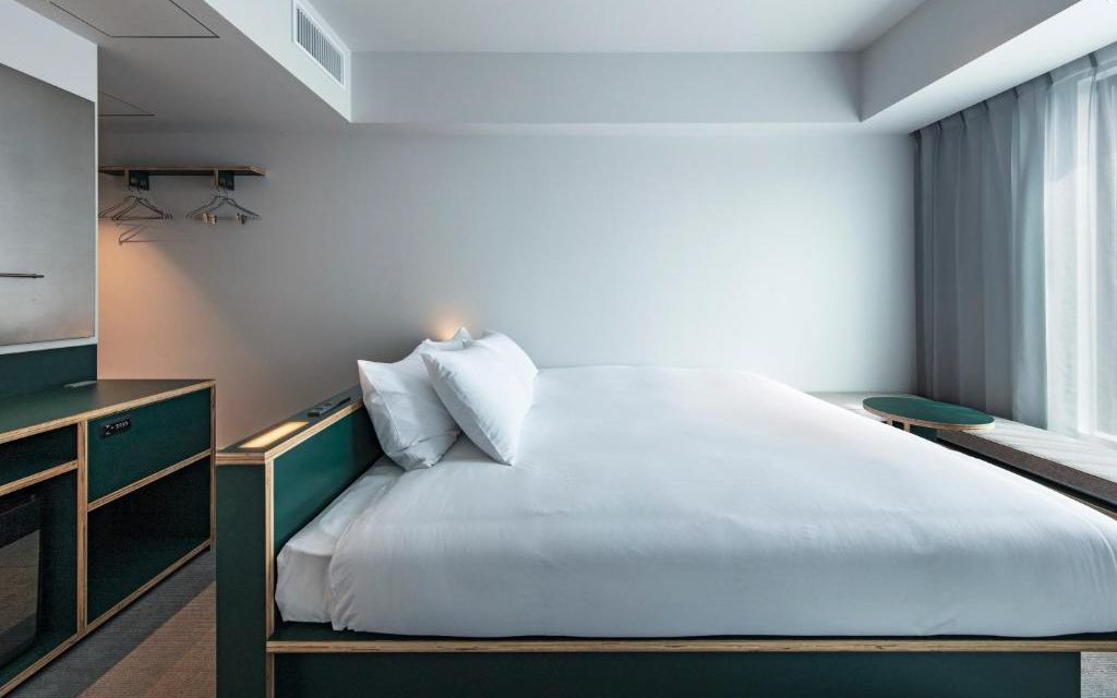 Hotel room with white bed, green plywood headboard and desk, and curtained window