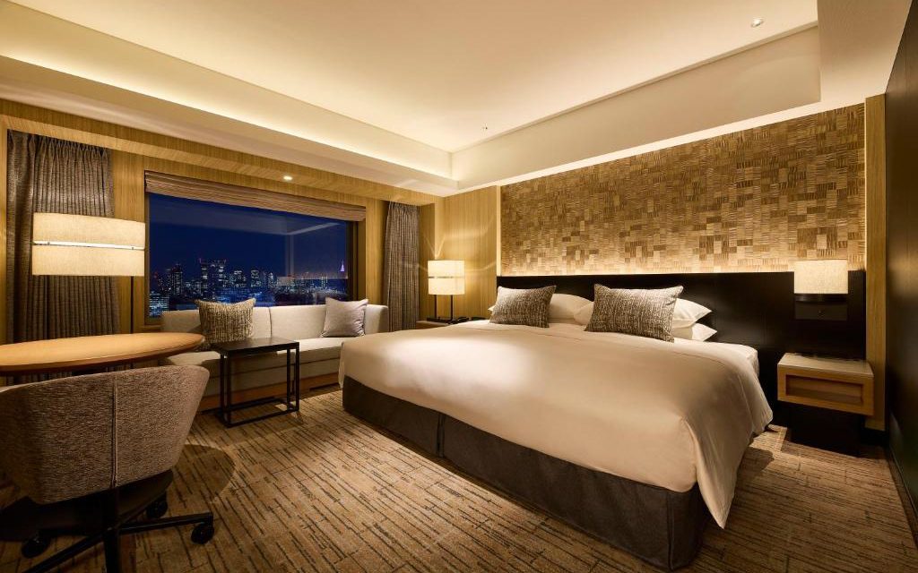 Hotel room with king bed, textured wood headboard, and city skyline visible through the window at night