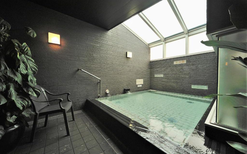 Indoor communal bath with dark stone tiling and glass ceiling at Dormy Inn Shibuya