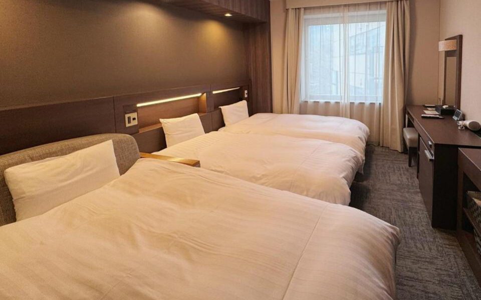 Hotel room with three made-up single beds, wooden headboard, and desk with TV