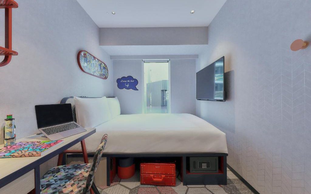Compact hotel room with double bed, work desk, wall-mounted TV, and in-room safe