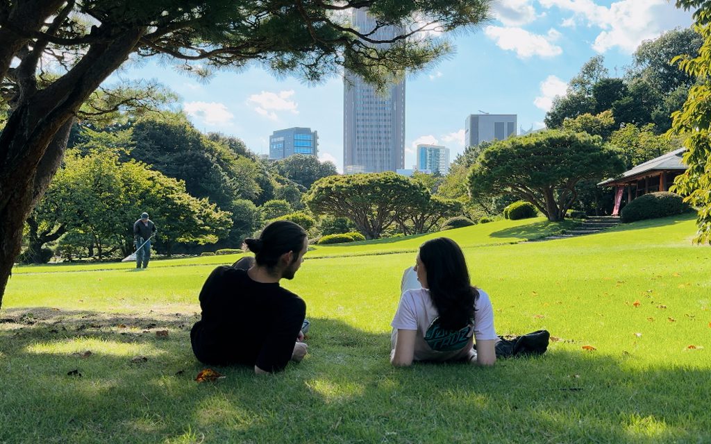 Couple sitting in the shade of a pine tree on the lawn at Shinjuku Gyoen, escaping the summer heat with the Tokyo skyline behind them