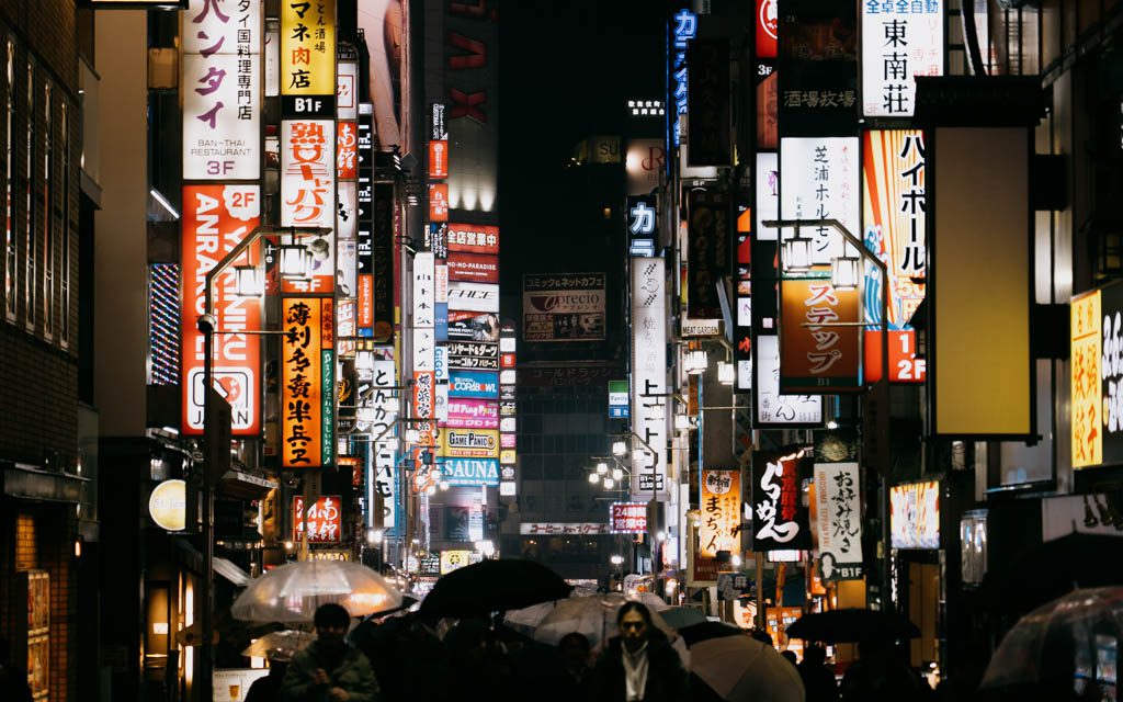 Towering neon signage above a crowded Shinjuku street at night with umbrellas