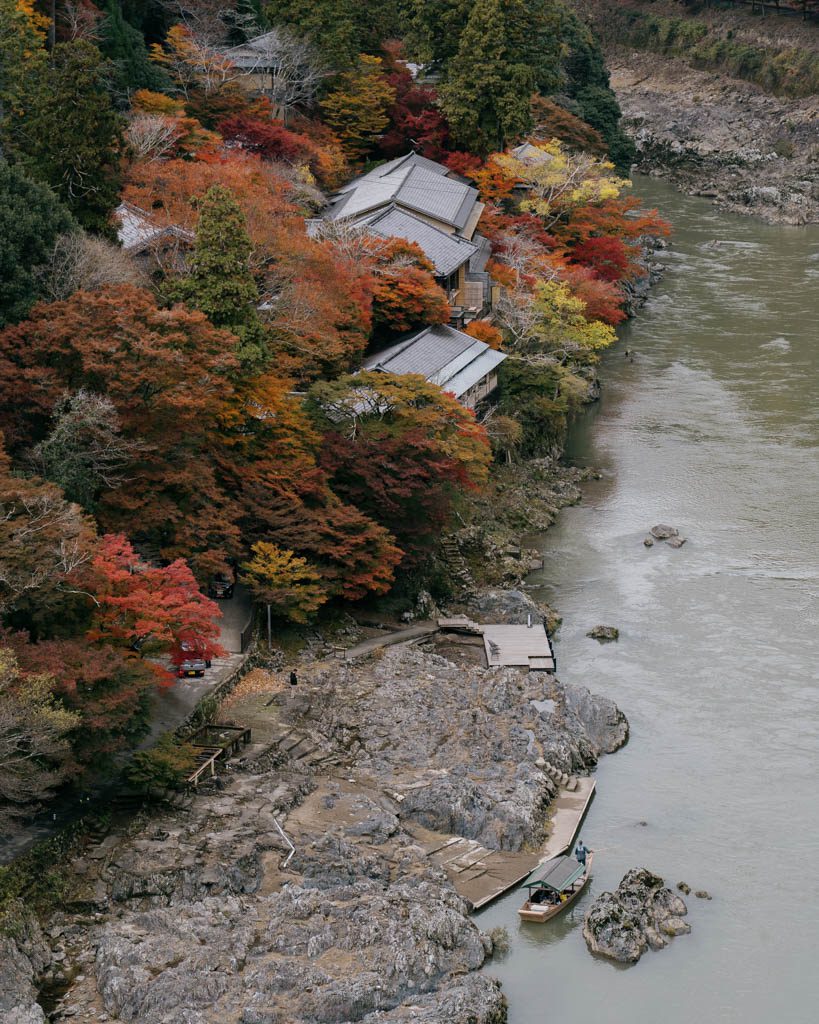 Traditional building surrounded by autumn foliage on the riverbank at Arashiyama, with a boat moored below
