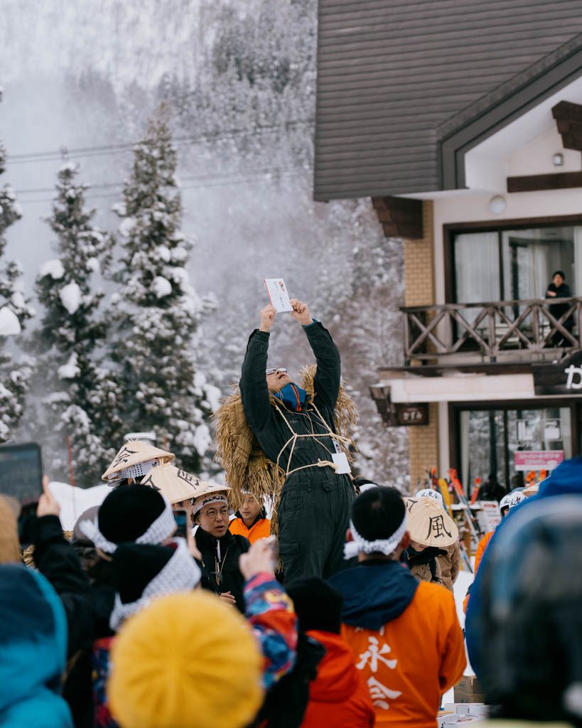Festival participant in a straw cape and hat holding a donation envelope above his head while a crowd looks on at the Dosojin Fire Festival
