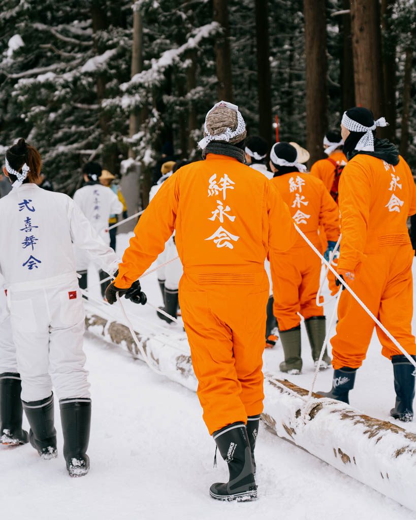 Participants in orange and white jumpsuits hauling a snow-covered log with ropes through the forest during the Dosojin Fire Festival