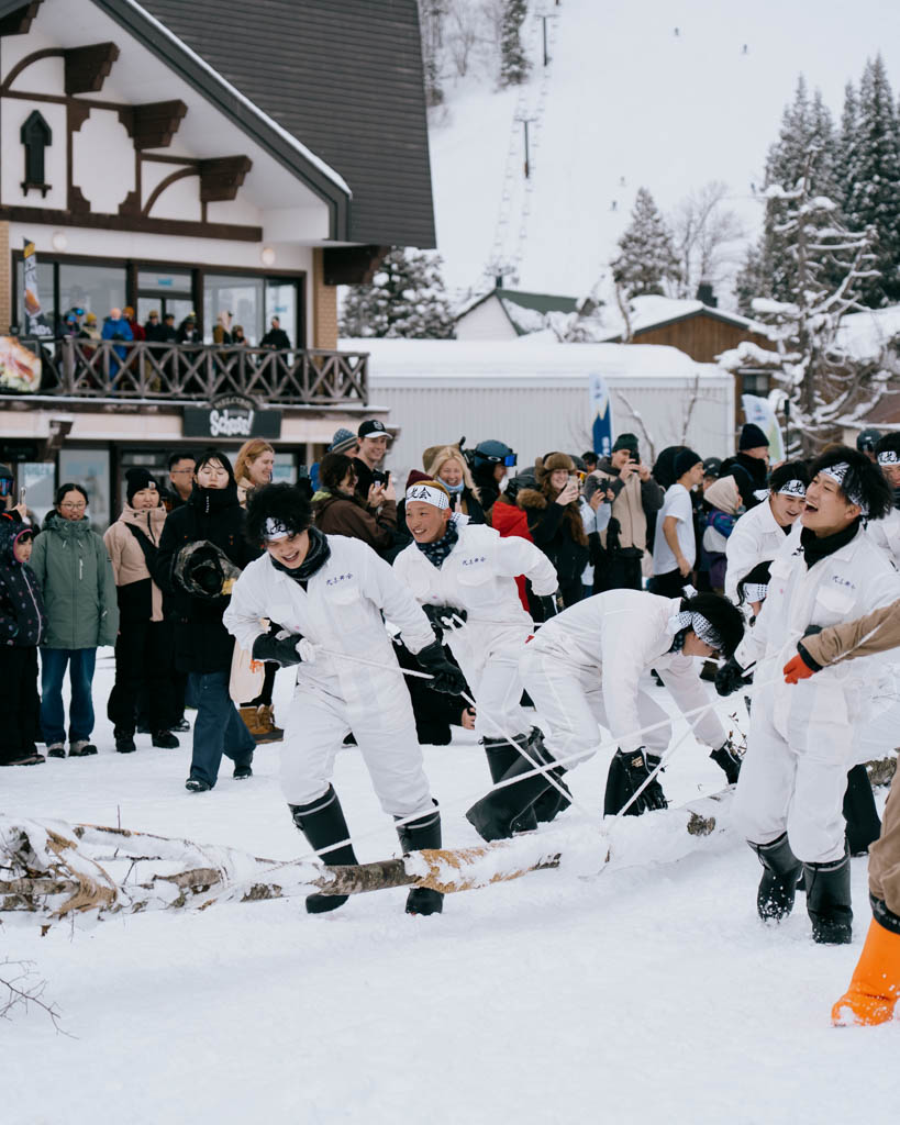Participants in white jumpsuits and headbands pulling a large log through the snow on ropes at the Dosojin Fire Festival, watched by a crowd