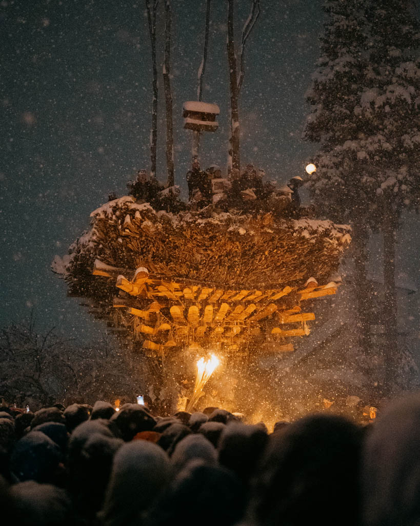 The wooden shaden pyre lit from below at night in heavy snow, with men gathered on top and a large crowd watching at the Dosojin Fire Festival