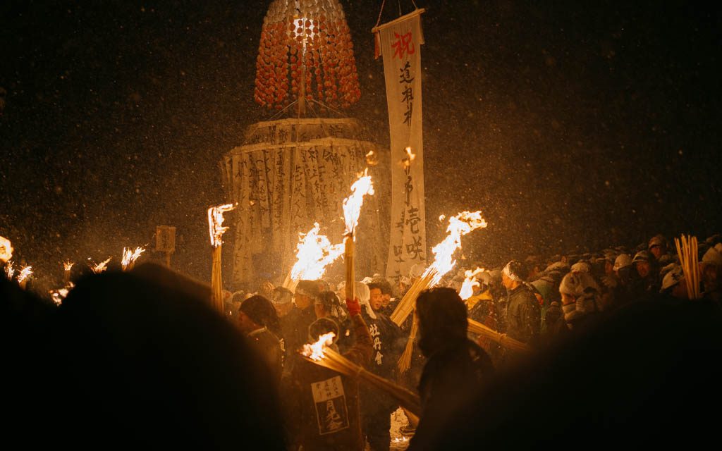 Villagers gathered in falling snow at night at the Dosojin Fire Festival, with unlit bundles of straw torches raised above the crowd