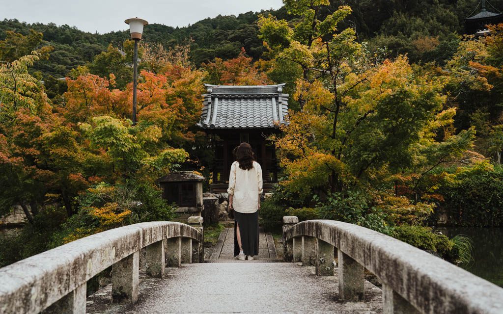 Woman walking across a stone bridge towards a small temple building at Eikan-do