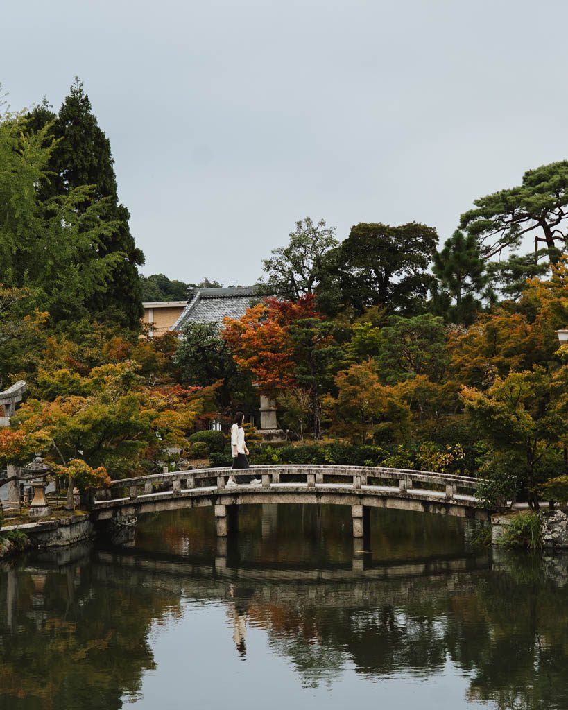 Woman crossing a stone bridge over a pond surrounded by autumn leaves at Eikan-do