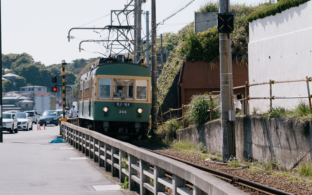 Green Enoden train traveling beside the coastal road in Kamakura with cars, power lines, and a red traffic light.