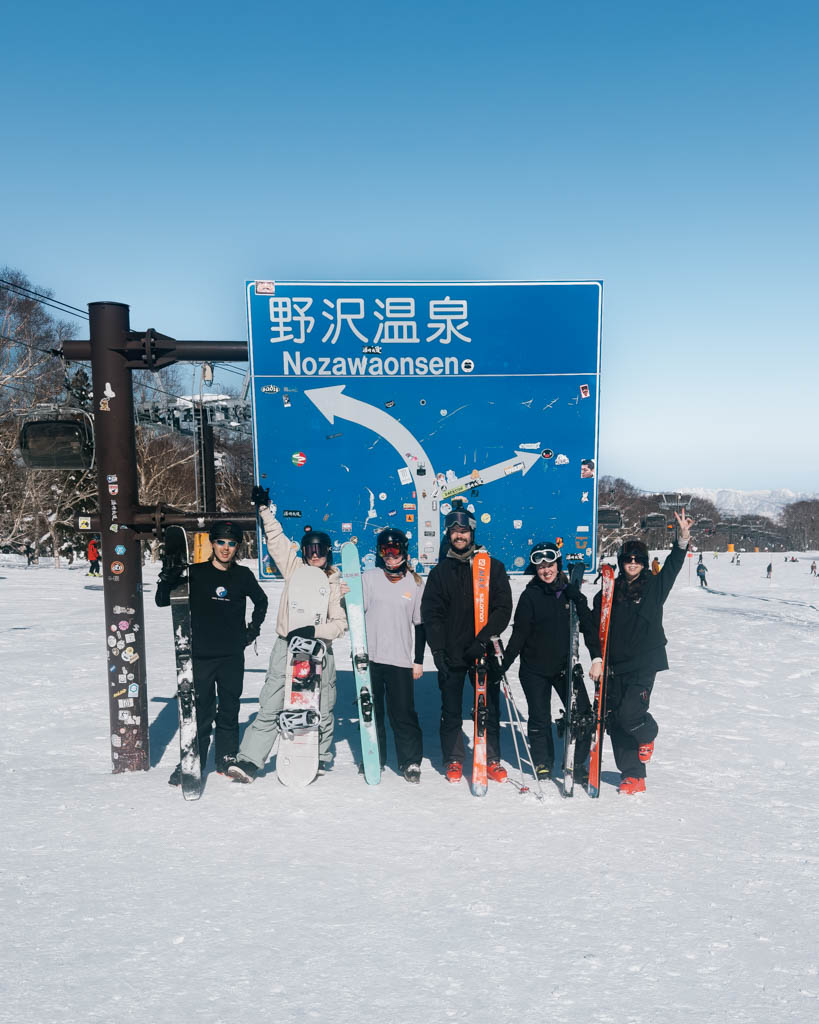 Group of skiers and snowboarders posing by the Nozawaonsen trail sign on a sunny day