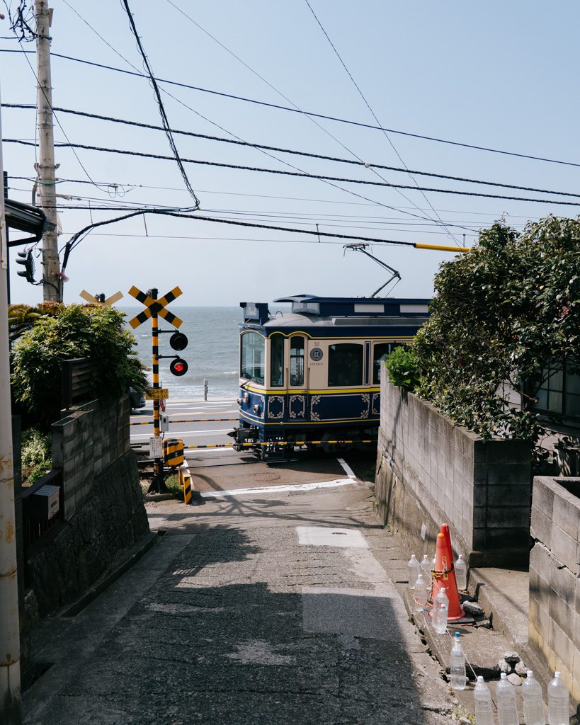 A vintage-style Enoden tram approaching a railway crossing at the end of a narrow Kamakura side street, with the ocean visible beyond.