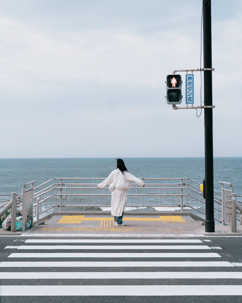 A visitor standing at a pedestrian crossing overlooking the ocean near Kamakurakōkō-mae Station.