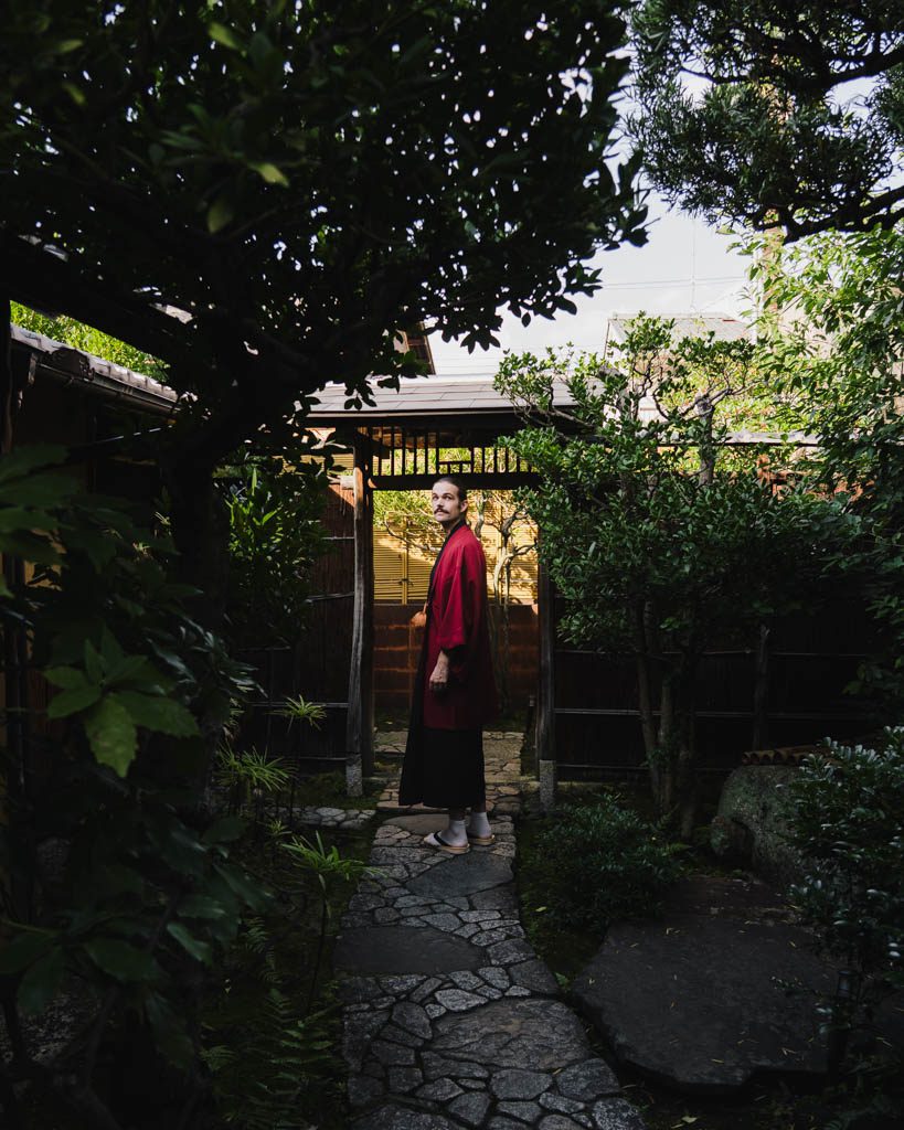 Man in a red kimono standing at a traditional wooden garden gate in Kyoto