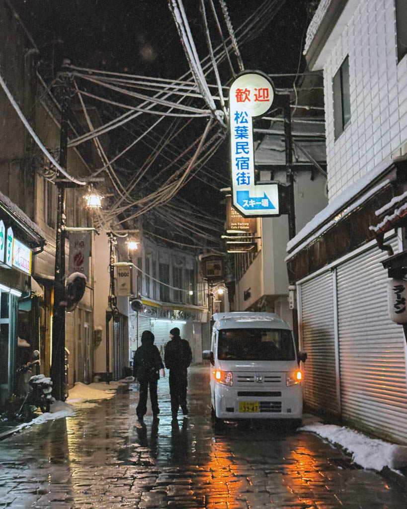 Snowy village street at night in Nozawa Onsen with a parked kei van and illuminated signage