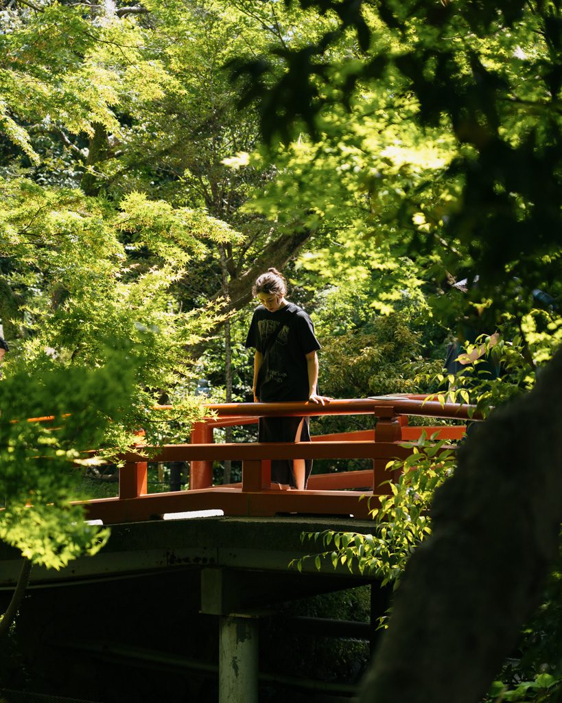 A red wooden bridge in a shaded garden at Tsurugaoka Hachiman-gu, with a visitor pausing to look over the railing.