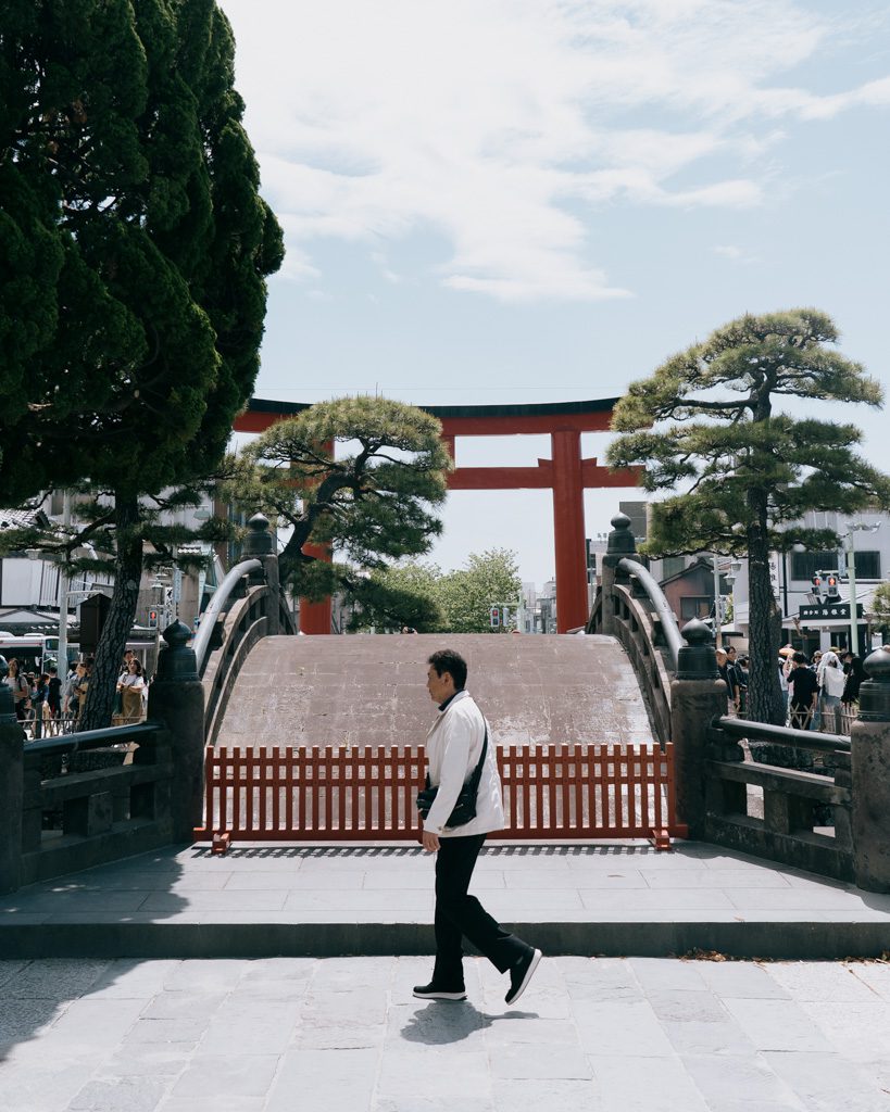 A large red torii gate and arched bridge at the entrance to Tsurugaoka Hachiman-gu shrine in Kamakura, with a visitor walking past.