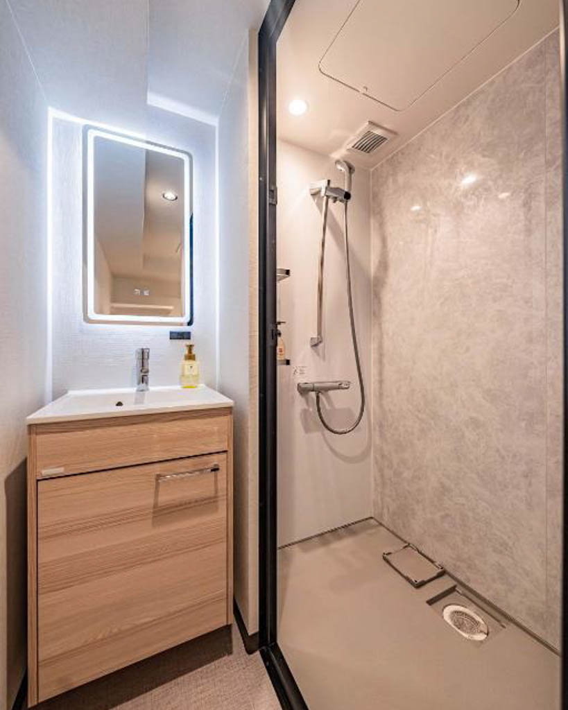 Compact hotel bathroom with a wooden vanity, lit mirror, and separate walk-in shower