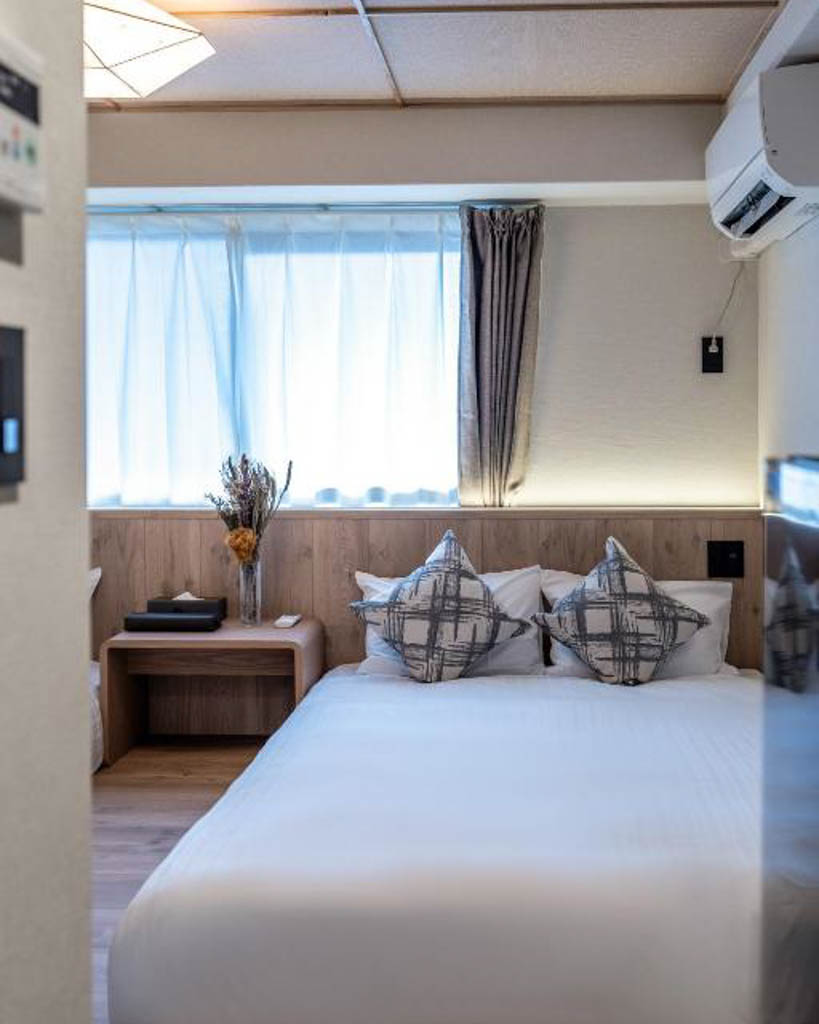 Hotel bedroom with a double bed, patterned cushions, bedside table with dried flowers, and curtained window