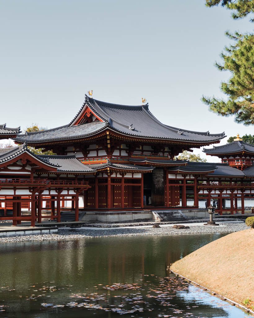 The red and black Phoenix Hall of Byodo-in temple in Uji, with gold phoenix statues on its roof, beside a pond on a clear day